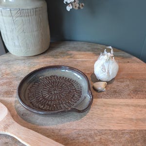 May include: A handmade ceramic garlic grater with a brown and tan glaze. The grater has a circular pattern in the center and a small handle. A bulb of garlic and a clove of garlic are next to the grater on a wooden surface.