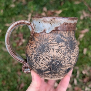 May include: Handmade ceramic mug with a brown and tan floral design. The mug features a large handle and a light blue and brown rim. The body of the mug is decorated with a sunflower pattern.