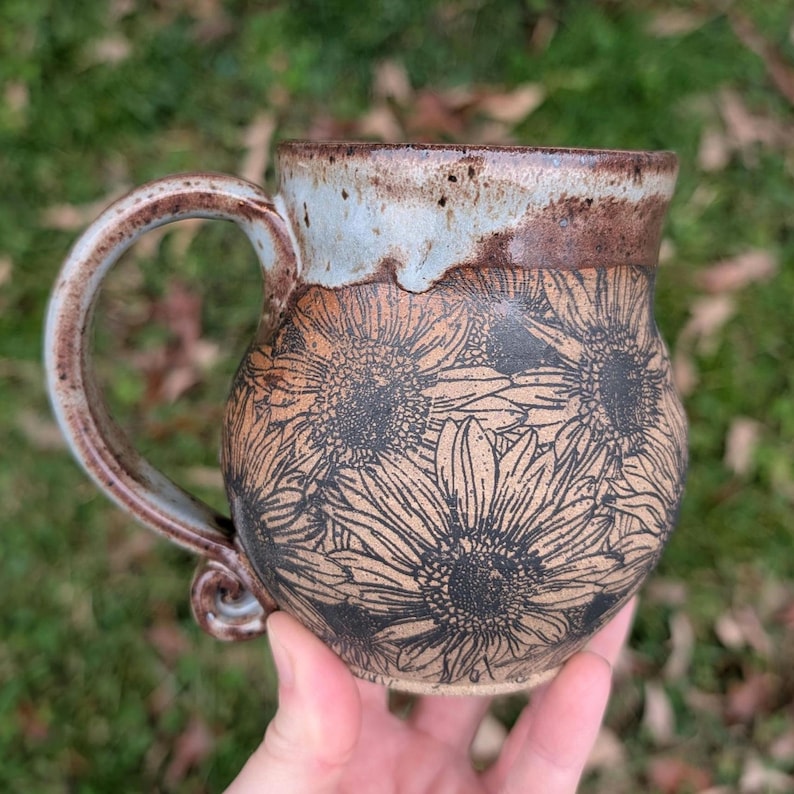 May include: Handmade ceramic mug with a brown and tan floral design. The mug features a large handle and a light blue and brown rim. The body of the mug is decorated with a sunflower pattern.