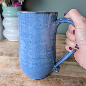 Op de afbeelding: Een handgemaakte, blauwe keramische mok met een grote handgreep. De mok heeft een textuuroppervlak en een donkerbruine rand. De mok wordt door een hand vastgehouden. Een vaas met bloemen staat op de achtergrond.