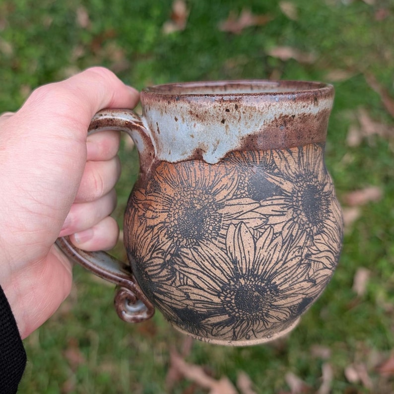 May include: A handmade ceramic mug with a sunflower design. The mug features a brown base with black sunflower illustrations, a light blue rim, and a brown handle. The mug is held in a hand against a blurred green background.