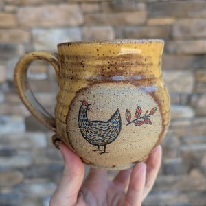Puede incluir: Taza de cerámica hecha a mano con un esmalte moteado marrón y beige. La taza presenta una ilustración dibujada a mano de una gallina con detalles azules y negros, y una ramita de hojas rojas. La taza tiene un asa grande y una forma redondeada.