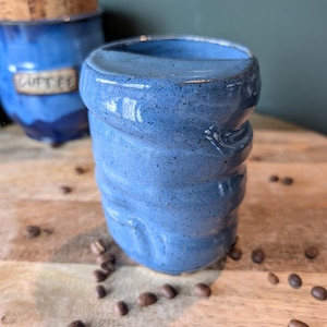 May include: A handmade, blue ceramic coffee mug with a textured, swirled design. The mug is surrounded by coffee beans on a wooden surface. In the background, a blue ceramic container with a cork lid is visible.