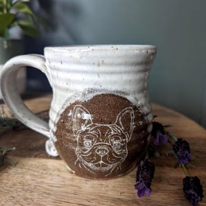 Puede incluir: Taza de cerámica hecha a mano con un acabado moteado blanco y marrón. La taza presenta una ilustración blanca detallada de la cara de un bulldog francés sobre un fondo marrón. La taza tiene un asa grande.