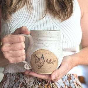 May include: A speckled white ceramic mug with a rounded body and a curved handle. The front features a circular tan-colored patch with a black line drawing of a chicken and a small branch with red leaves. The mug has a textured, rustic appearance.