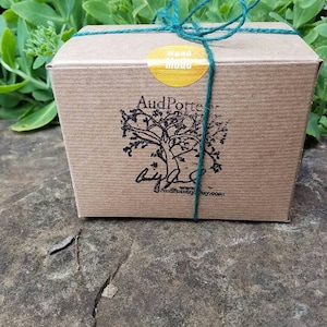 May include: A small, brown cardboard box tied with teal twine. A yellow sticker with the words "Hand Made" is on the top. The box has a tree logo and the text "AudPorter" printed on the front.