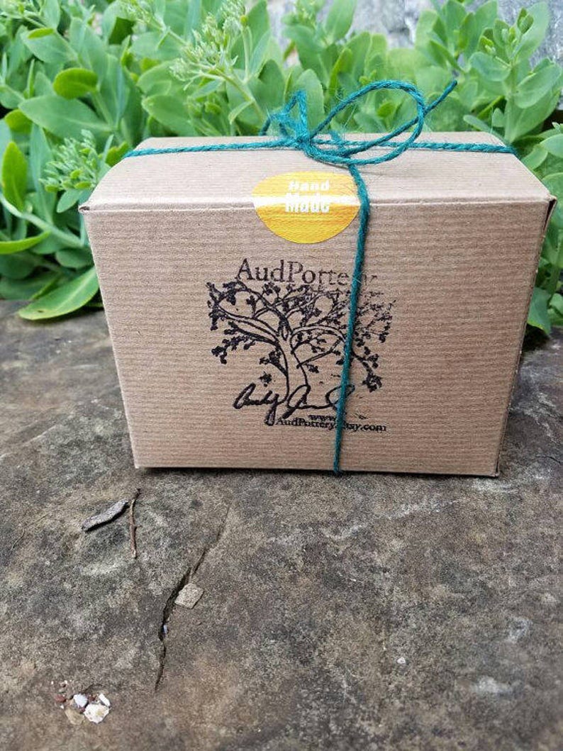 May include: A small, brown cardboard box tied with teal twine. A yellow sticker with the words "Hand Made" is on the top. The box has a tree logo and the text "AudPorter" printed on the front.