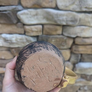 May include: A handmade ceramic mug with a yellow handle and a floral design in black. The bottom of the mug has a handwritten signature. The mug is held against a stone wall background.