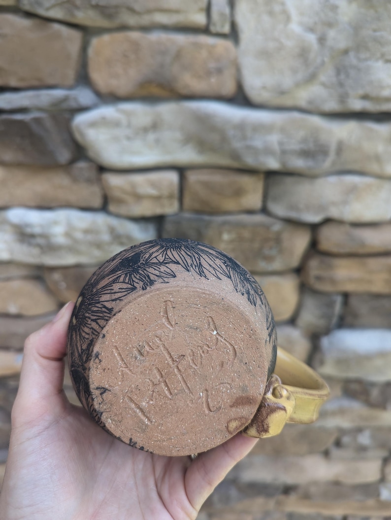 May include: A handmade ceramic mug with a yellow handle and a floral design in black. The bottom of the mug has a handwritten signature. The mug is held against a stone wall background.