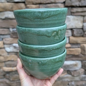 Op de afbeelding: Een stapel van vier handgemaakte keramische kommen met een gedempte groene glazuur. De kommen hebben een onregelmatige vorm en een getextureerd oppervlak. Elke kom heeft een diameter van ongeveer 12,7 cm en is 5 cm hoog.