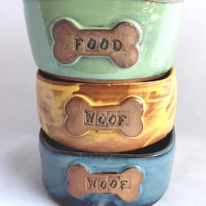 Puede incluir: Una pila de tres cuencos de cerámica para perros en tonos verde, amarillo y azul. Cada cuenco tiene un diseño en forma de hueso con las palabras "FOOD" o "WOOF". Los cuencos están diseñados para comida de mascotas.
