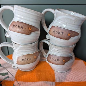 May include: Four stacked, handmade ceramic mugs. Each mug is white with a brown, state-shaped design that reads "HOME" and features a small heart. The mugs have large handles and a rustic, textured appearance.