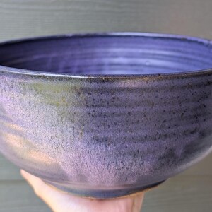 May include: A deep, round ceramic bowl featuring a variegated glaze that transitions from dark purple to lighter violet tones. The exterior surface displays subtle horizontal ridges and a speckled texture, with a smooth, dark interior finish.