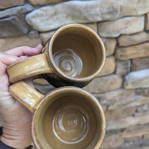 May include: Two handmade ceramic mugs with a tan glaze and dark brown rims. The mugs have a rounded shape with a handle and a textured exterior. The interior of the mugs is a lighter tan color. The mugs are likely for coffee or tea.