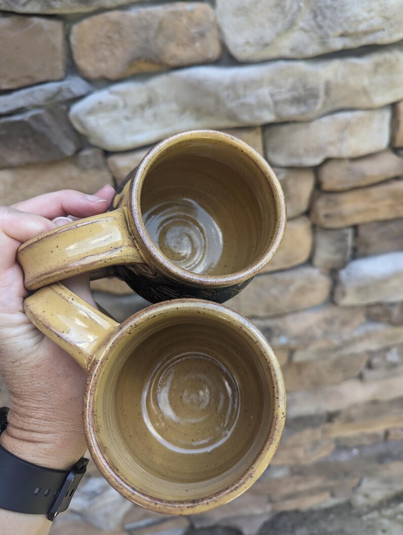 May include: Two handmade ceramic mugs with a tan glaze and dark brown rims. The mugs have a rounded shape with a handle and a textured exterior. The interior of the mugs is a lighter tan color. The mugs are likely for coffee or tea.