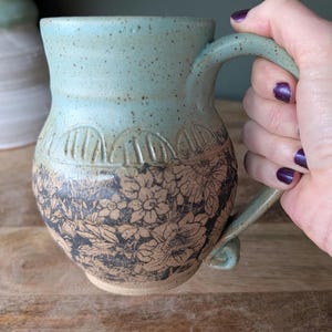 Puede incluir: Una taza de cerámica hecha a mano con un esmalte azul claro moteado. La mitad inferior presenta un detallado patrón floral en negro y beige. La taza tiene un asa curva y está sostenida por una persona.