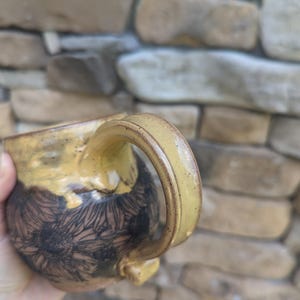May include: A handmade ceramic mug with a yellow handle and rim. The mug's body is decorated with a black floral pattern on a brown background. The mug is held in a hand, with a stone wall in the background.