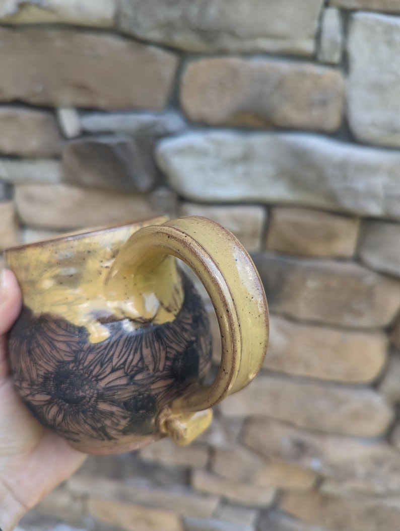 May include: A handmade ceramic mug with a yellow handle and rim. The mug's body is decorated with a black floral pattern on a brown background. The mug is held in a hand, with a stone wall in the background.