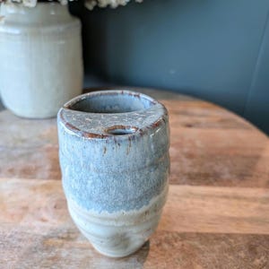 Può includere: Un piccolo vaso in ceramica fatto a mano con una forma unica. Il vaso è principalmente azzurro con una superficie strutturata e un bordo marrone. La parte inferiore è di colore beige chiaro. Il vaso è alto circa 10 cm.