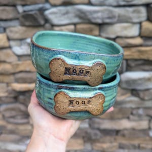 May include: Two turquoise ceramic dog bowls stacked on top of each other. Each bowl has a bone-shaped accent with the word "WOOF" in the center. The bowls have a rustic, handmade appearance, with visible texture and color variations.