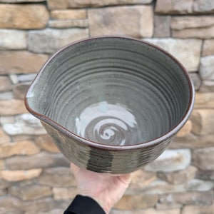 May include: A handmade ceramic mixing bowl with a spout, featuring a gray-green glaze and a spiral design on the interior. The bowl has a dark brown rim and is held by a person.