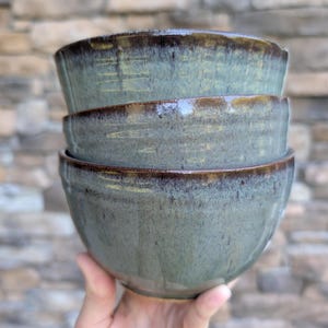 May include: A stack of three ceramic bowls featuring a mottled sage green glaze with dark brown, metallic-toned rims. The bowls have a smooth, rounded shape with subtle vertical texture visible beneath the glossy, multi-tonal finish.