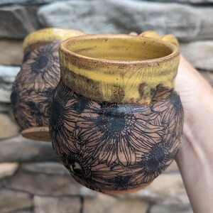May include: Two handmade ceramic mugs. The mugs are brown with a yellow rim and handle. The exterior of the mugs are decorated with a black sunflower design. The mugs are held in front of a stone background.