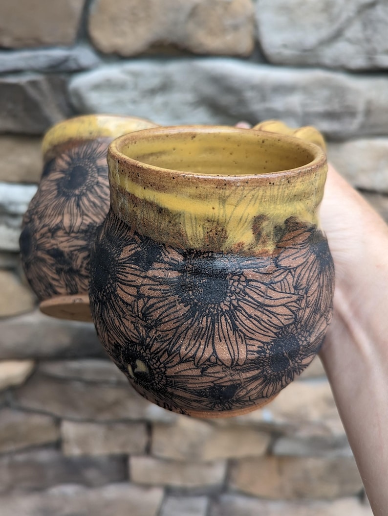 May include: Two handmade ceramic mugs. The mugs are brown with a yellow rim and handle. The exterior of the mugs are decorated with a black sunflower design. The mugs are held in front of a stone background.