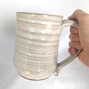May include: A tall, cylindrical ceramic mug with a light cream-colored glaze and a visible vertical crack running down the side. The mug features a textured, ribbed surface with small dark speckles throughout and a sturdy, curved handle.