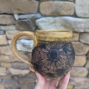 May include: A handmade ceramic mug with a yellow rim and handle. The body of the mug is brown with a black sunflower design. The mug is held in a person's hand against a stone background.