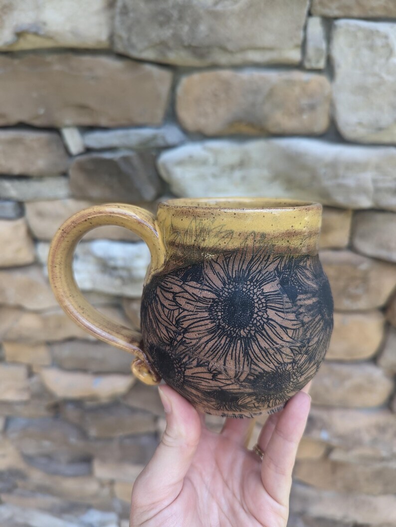 May include: A handmade ceramic mug with a yellow rim and handle. The body of the mug is brown with a black sunflower design. The mug is held in a person's hand against a stone background.