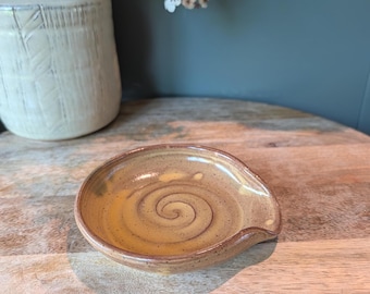 Pode incluir: Um descanso de colher de cerâmica feito à mão com um design em espiral em tons de castanho e amarelo. O descanso de colher tem uma forma curva com um pequeno lábio. Um vaso bege e flores brancas no fundo.