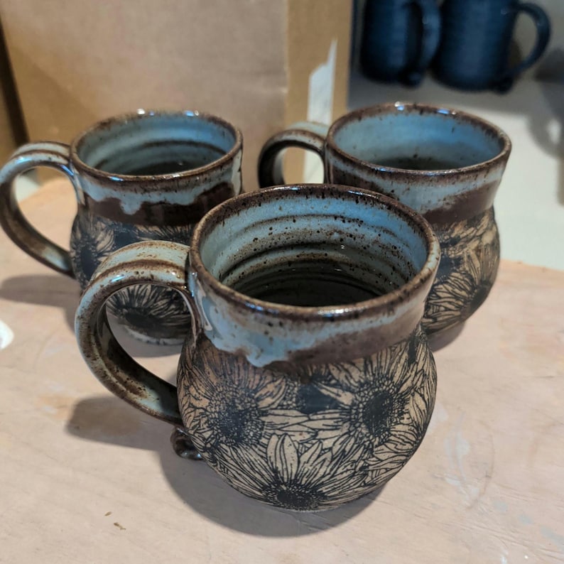 May include: Three handmade ceramic mugs with sunflower designs. The mugs have a brown base, a light blue rim, and a dark brown handle. The sunflower design is etched into the ceramic. The mugs are on a light brown surface.