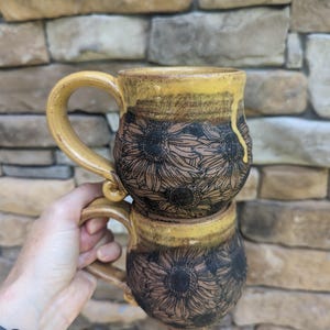 May include: Two ceramic mugs stacked, featuring a sunflower design. The mugs have a yellow rim and handle, with a brown base. The sunflower design is etched in black. The mugs are held in front of a stone wall.