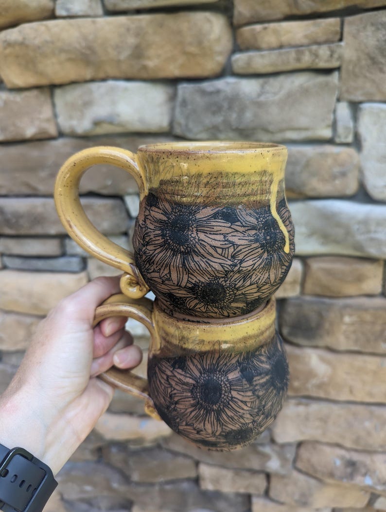 May include: Two ceramic mugs stacked, featuring a sunflower design. The mugs have a yellow rim and handle, with a brown base. The sunflower design is etched in black. The mugs are held in front of a stone wall.