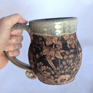 Puede incluir: Una taza de cerámica hecha a mano con un diseño floral. La taza tiene una base marrón con patrones florales negros y un borde y asa verde claro. La taza está sostenida por una mano.