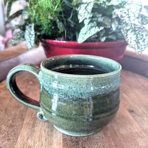 May include: A handmade ceramic mug with a green glaze. The mug has a rounded shape, a handle, and a small foot. The interior of the mug is filled with a dark liquid. A red pot with green plants is in the background.