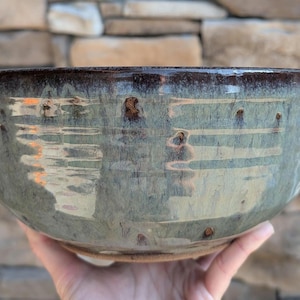 May include: A handmade ceramic bowl with a wide, rounded shape. The bowl features a glossy, mottled glaze in shades of blue-green and brown. The rim is a darker brown, and the bowl has a rustic, organic feel.