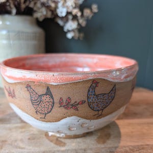 May include: A handmade ceramic bowl with a rustic design. The bowl features a coral-colored interior and a brown exterior with hand-painted chickens and red leaf accents. The base is white with small holes.