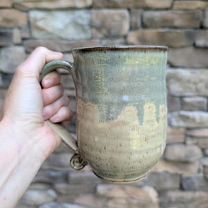 Peut inclure: Une tasse en céramique avec un vernis bicolore, présentant une partie supérieure vert sauge et une partie inférieure beige mouchetée. La tasse possède une anse robuste avec un petit détail décoratif enroulé à la base et un corps cylindrique.
