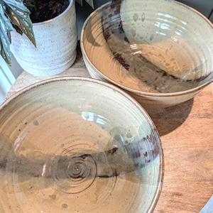 Op de afbeelding: Twee handgemaakte keramische kommen met een beige en bruin glazuur. De kommen hebben een breed, ondiep ontwerp en staan op een houten oppervlak. De kommen staan naast een witte keramische pot met een groene plant.