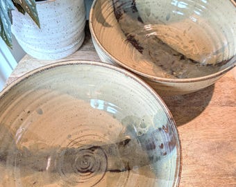 May include: Two handmade ceramic bowls with a beige and brown glaze. The bowls have a wide, shallow design and are placed on a wooden surface. The bowls are next to a white ceramic pot with a green plant.