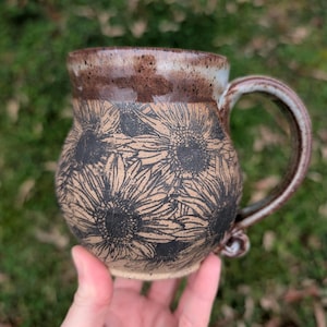 May include: A handmade ceramic mug with a sunflower design. The mug features a tan base with black sunflower illustrations and a brown rim and handle. The mug is designed for hot or cold beverages.