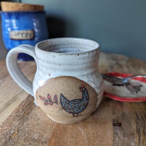May include: A speckled white ceramic mug with a rounded body and a handle. The front features a circular tan patch with a hand-painted blue chicken and a small red leaf sprig. The mug has a textured, slightly uneven surface with visible speckles.