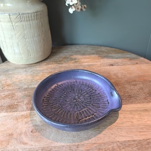 May include: A small, round, purple ceramic spoon rest with a textured, circular pattern in the centre. The spoon rest has a small lip on one side. A cream-coloured vase is in the background.