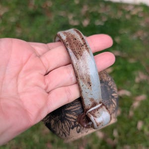May include: A ceramic mug with a sunflower design. The mug has a light blue handle with brown spots. The body of the mug is tan with a black sunflower design. The mug is held in a hand.