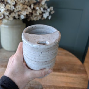 May include: A speckled, off-white ceramic utensil holder with a curved top and a small hole. The cylindrical holder has a textured, ridged design and is held by a person's hand. The background includes a vase with flowers and a wooden table.