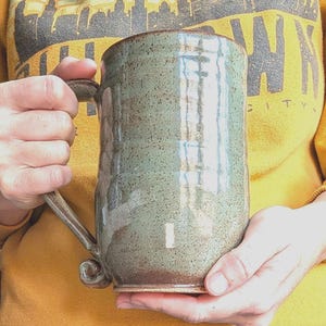 Könnte beinhalten: Eine handgefertigte, gesprenkelte Keramiktasse in Grün- und Brauntönen. Die Tasse hat einen großen, gebogenen Henkel und eine leicht unregelmäßige Form, was darauf hindeutet, dass sie handgefertigt ist. Die Tasse wird von einer Person gehalten, die einen gelben Pullover trägt.