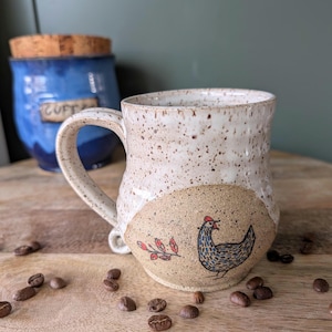 Könnte beinhalten: Eine gesprenkelte Keramiktasse mit Henkel, mit einer handgezeichneten Hühnerillustration. Die Tasse hat einen beigefarbenen Boden und einen weißen oberen Teil. Ein blaues Glas mit Korkdeckel befindet sich im Hintergrund.