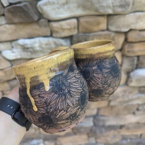 May include: Two ceramic mugs with a sunflower design. The mugs have a brown base with black sunflower patterns and a yellow rim. The glaze drips down the side of one mug. The background is a stone wall.